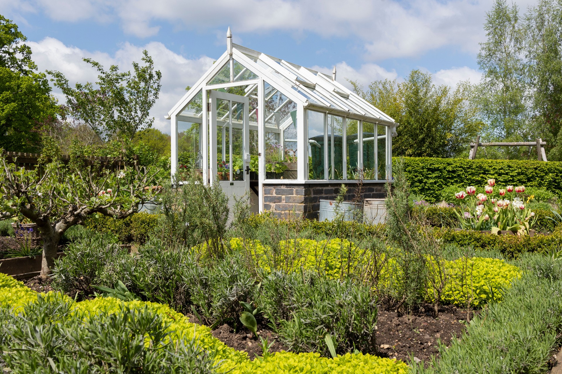 Gemüsebeet mit Hochbeeten und einem modernen Gewächshaus im blühenden Garten